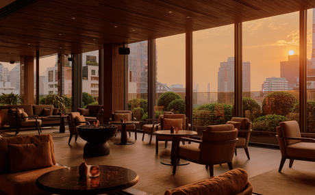 Aman Nai Lert Bangkok lounge with floor-to-ceiling windows overlooking Bangkok at sunset, warm lighting and contemporary furnishings.