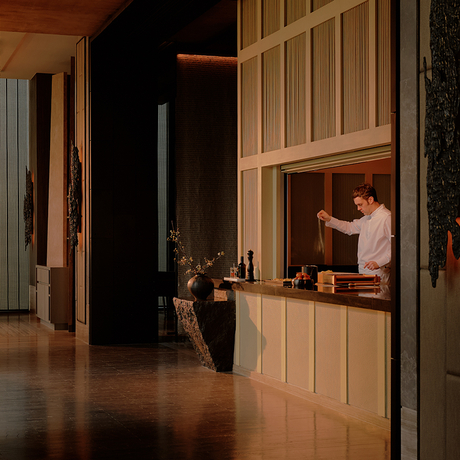 Two staff members work at the reception desk of Aman Nai Lert Bangkok, framed by tall wooden doors in a dimly lit corridor.