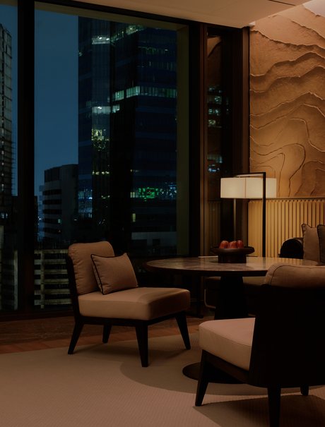 Mid-century modern armchair and side table in a Bangkok hotel room overlooking the city at dusk.