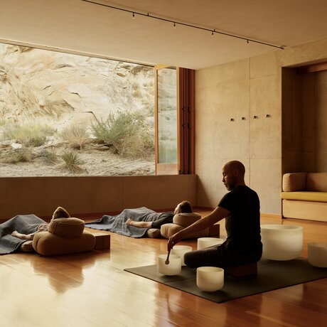 A person sits cross-legged on a wooden floor during a sound bowl wellness experience at Amangiri, with a large landscape artwork on the wall.