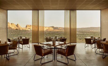 Amangiri restaurant with floor-to-ceiling windows overlooking desert landscape, featuring round dining tables and mid-century modern seating.