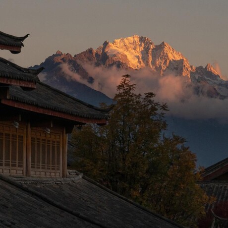Golden sunrise light illuminates snow-capped mountain peaks above Amandayan's roofline, with mist rising through forested valleys below.