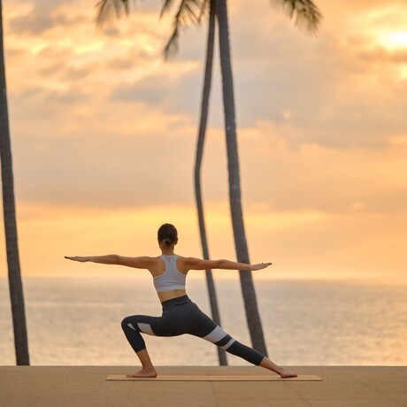 A person practises yoga at sunset by the sea at Amanpuri, Thailand, framed by tall palm trees.