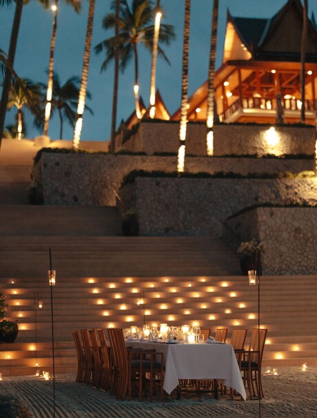 Beach barbecue dinner table setup at Amanpuri, Thailand, with candlelit pathway and illuminated dining pavilion at dusk.