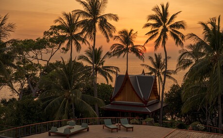 Amanpuri villa at sunset with palm trees silhouetted against an orange sky.