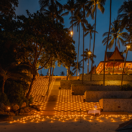 Candlelit courtyard at dusk, Amanpuri resort in Thailand, with warm lights casting patterns across stone pavement and palm trees silhouetted against twilight sky.