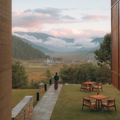 Outdoor dining area at Gangtey Lodge, Amankora, overlooking a misty valley with forested mountains and guests walking on the lawn.