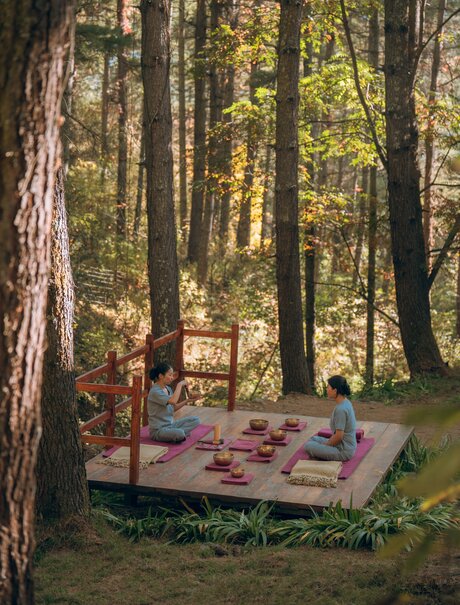 Woman practising sound bath meditation on wooden platform amongst tall forest trees at Amankora, Thimphu Lodge.