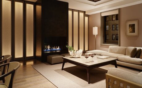 Evening view of a corner suite living room at Aman New York, featuring a fireplace, neutral tones, and contemporary furnishings.