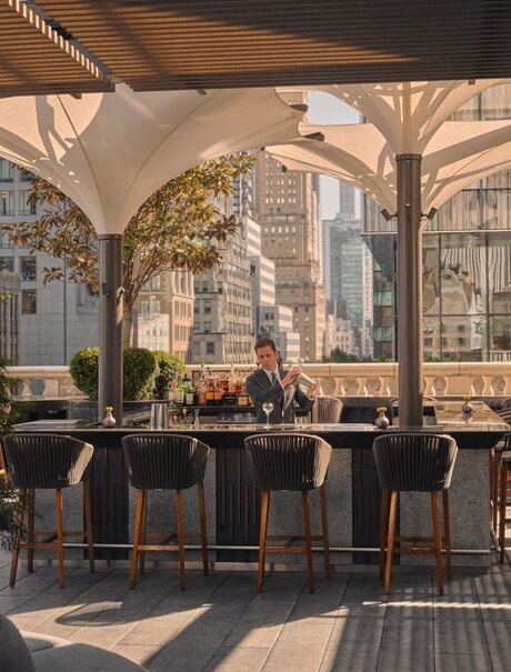 Bartender crafting cocktails at Aman New York's corner bar and garden terrace, shaded by canvas awnings.