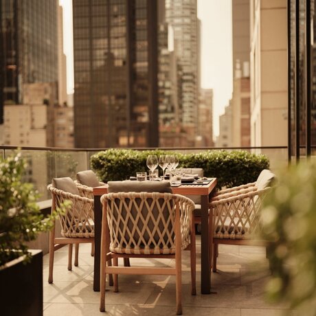Woven chair and table on a sunlit terrace at Aman New York, with city skyscrapers visible beyond.