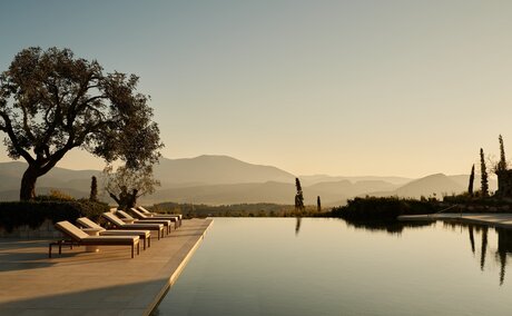 Sunbeds beside the main pool at Amanzoe resort, Greece, with cypress trees reflected in still water at dusk.