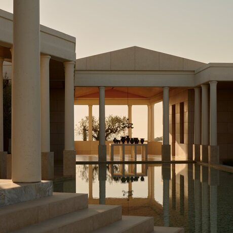 Amanzoe resort entrance pavilion at dusk, with illuminated wooden doors and columns.