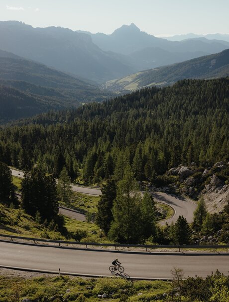 Cyclist on mountain road with alpine valley and forested peaks at Aman Rosa Alpina resort.