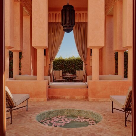 Symmetrical pavilion at Amanjena with terracotta walls, two chairs flanking a geometric mosaic floor, framing a view of the gardens beyond.