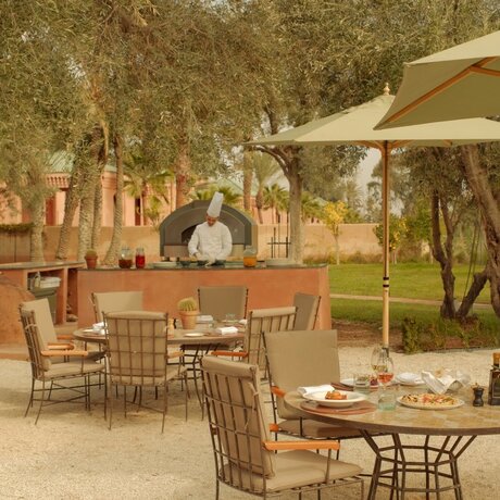Outdoor dining area at Amanjena with terracotta courtyard, olive trees, and shaded seating beneath cream parasols.
