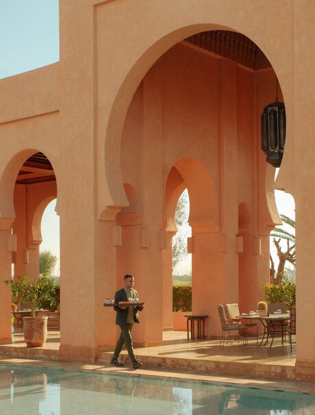 Terracotta arches frame the main pool at Amanjena, Morocco, with a figure walking across the tranquil water's edge.