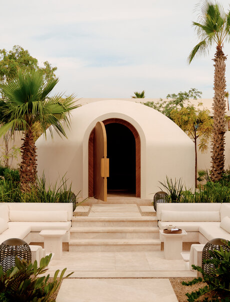 Amanvari spa entrance with curved white walls and wooden door, flanked by palm trees and stone steps in Mexico.
