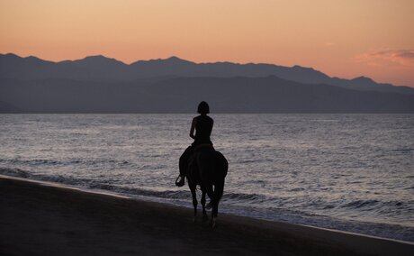 Horseback rider on a beach at sunset, mountains silhouetted across the water at Amanvari.