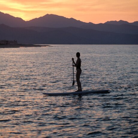 Silhouetted figure paddleboarding on calm waters at sunset, mountains beyond, at Amanvari, Mexico.