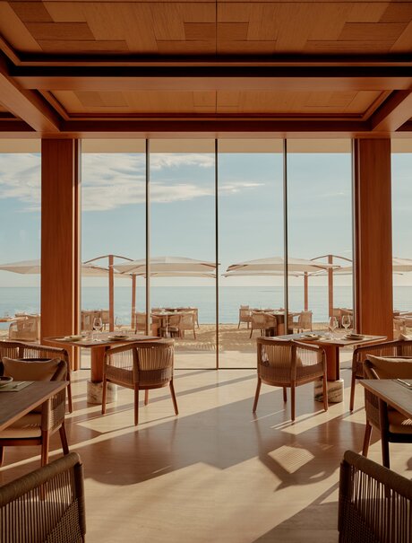 Amanvari dining venue with wooden beams, chairs, and expansive windows overlooking desert landscape.
