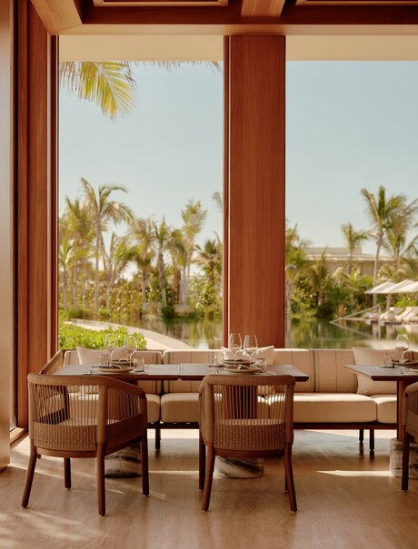 Amanvari dining area with wooden table and chairs overlooking palm-lined gardens through floor-to-ceiling windows.