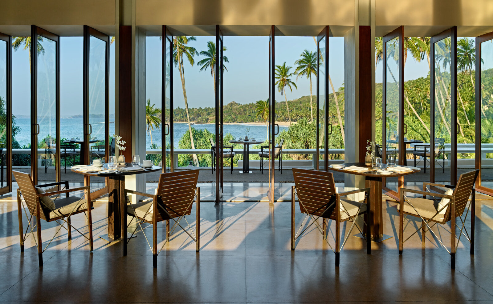 The restaurant laid for a meal, with glass walls opening onto the bay and palms