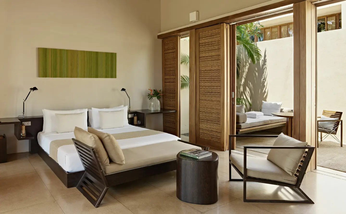 A suite bedroom with a low king bed, timber-framed armchairs and tall sliding doors open to the courtyard