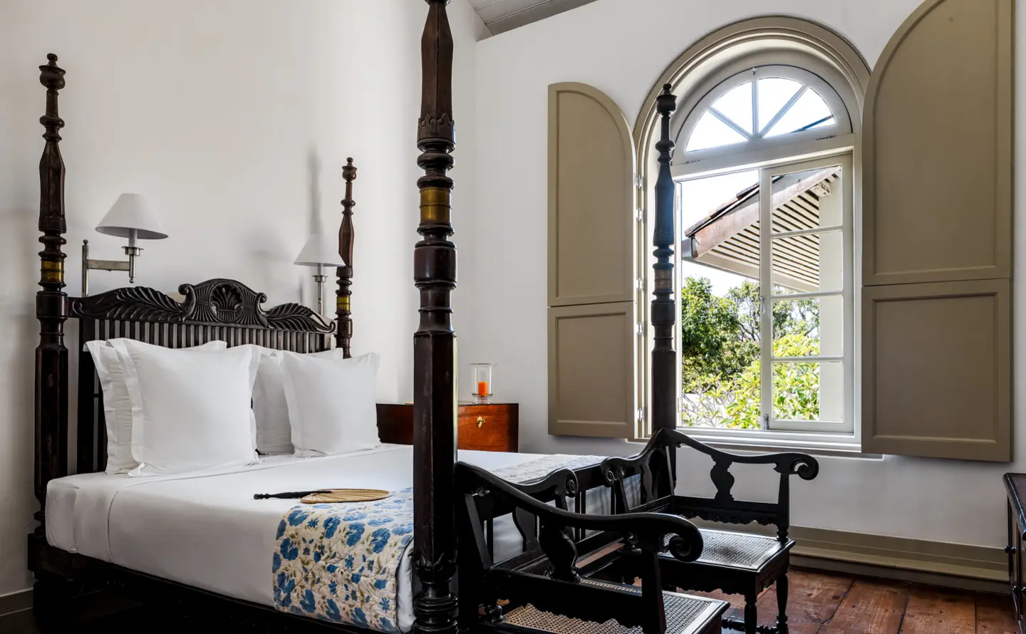 A bedroom with a dark carved four-poster bed, white linen and a tall arched window with its shutters open