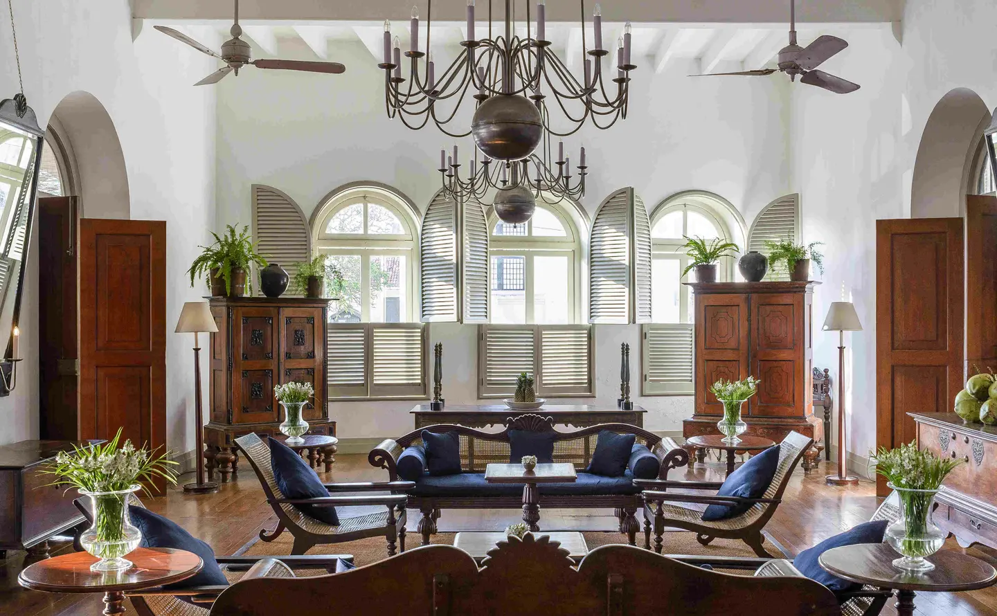 A high white lounge with a chandelier, ceiling fans, arched shuttered windows and sofas in navy upholstery