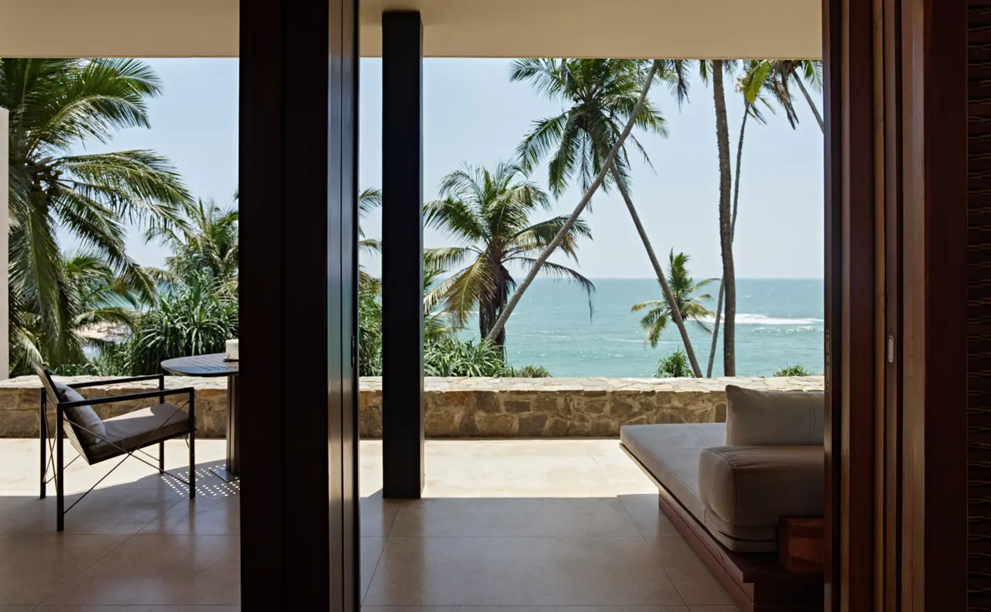A suite terrace with a daybed between dark pillars, looking out through palms to the surf
