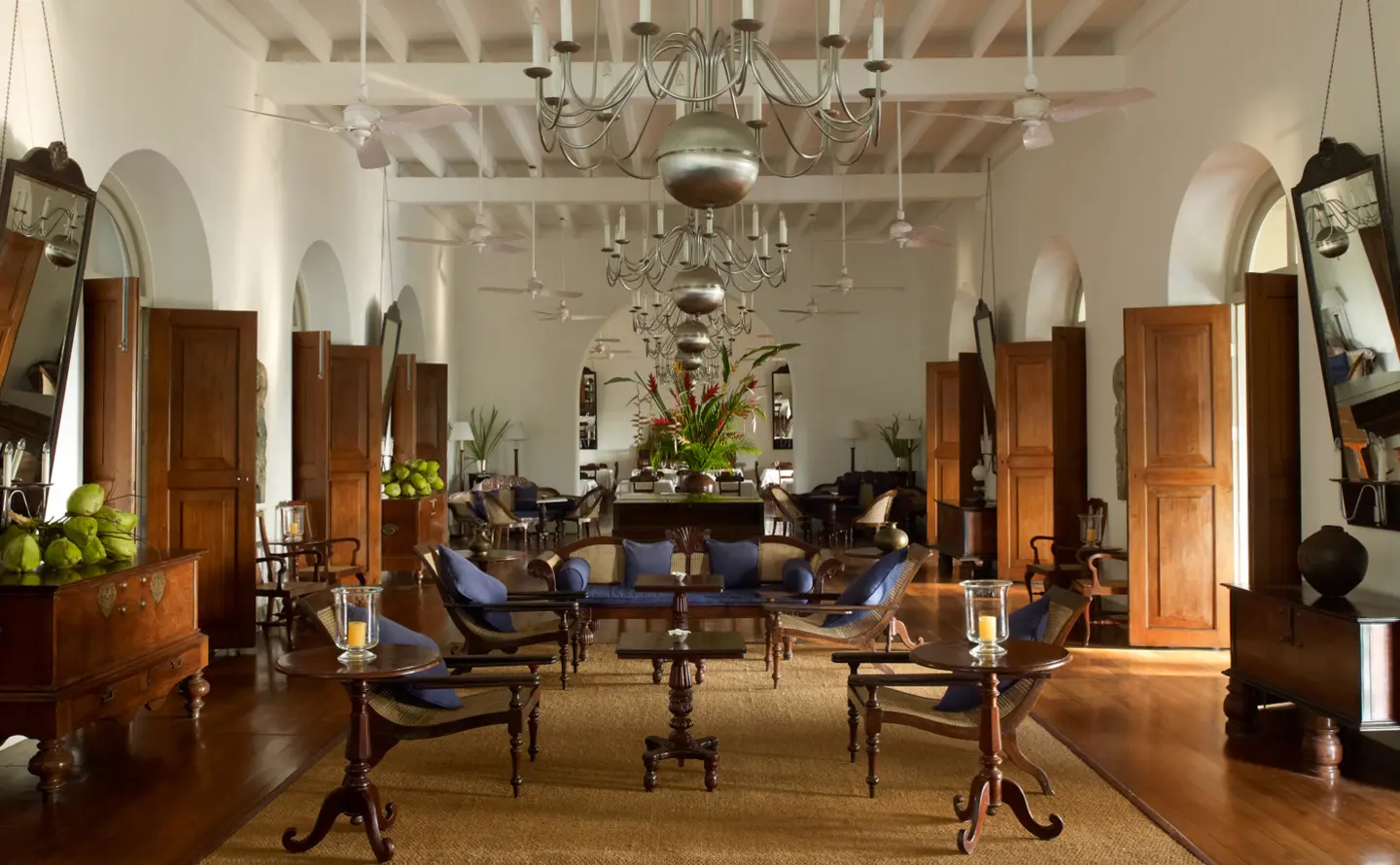 A long hall with chandeliers, tall wooden doors and armchairs grouped around low tables on a timber floor