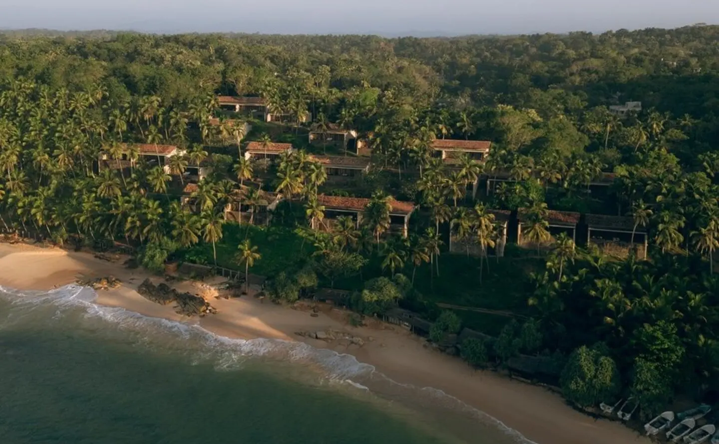 Amanwella from the air: tiled suite roofs scattered through coconut palms on a hillside above a curved sand beach and green sea