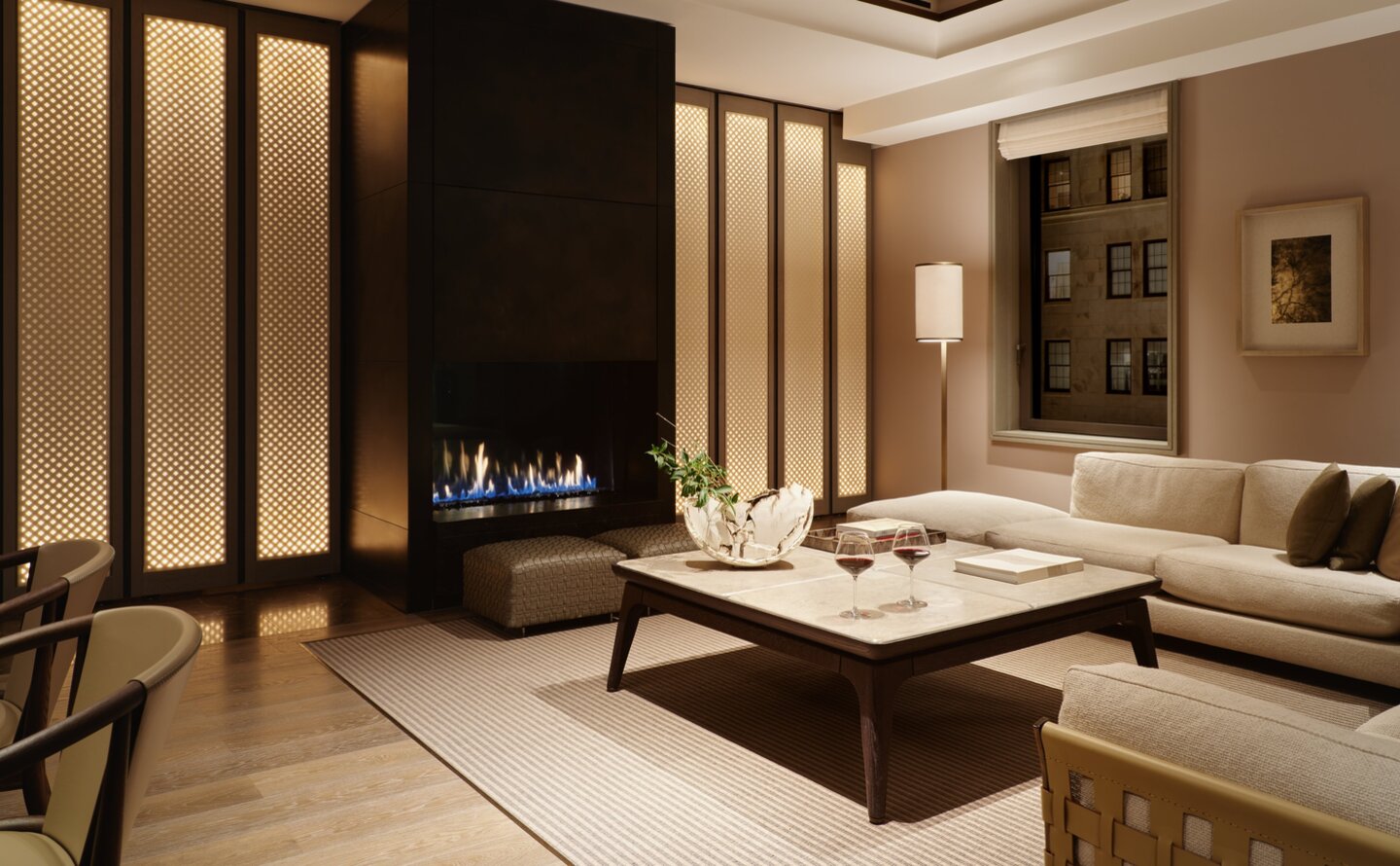Evening view of a corner suite living room at Aman New York, featuring a fireplace, neutral tones, and contemporary furnishings.