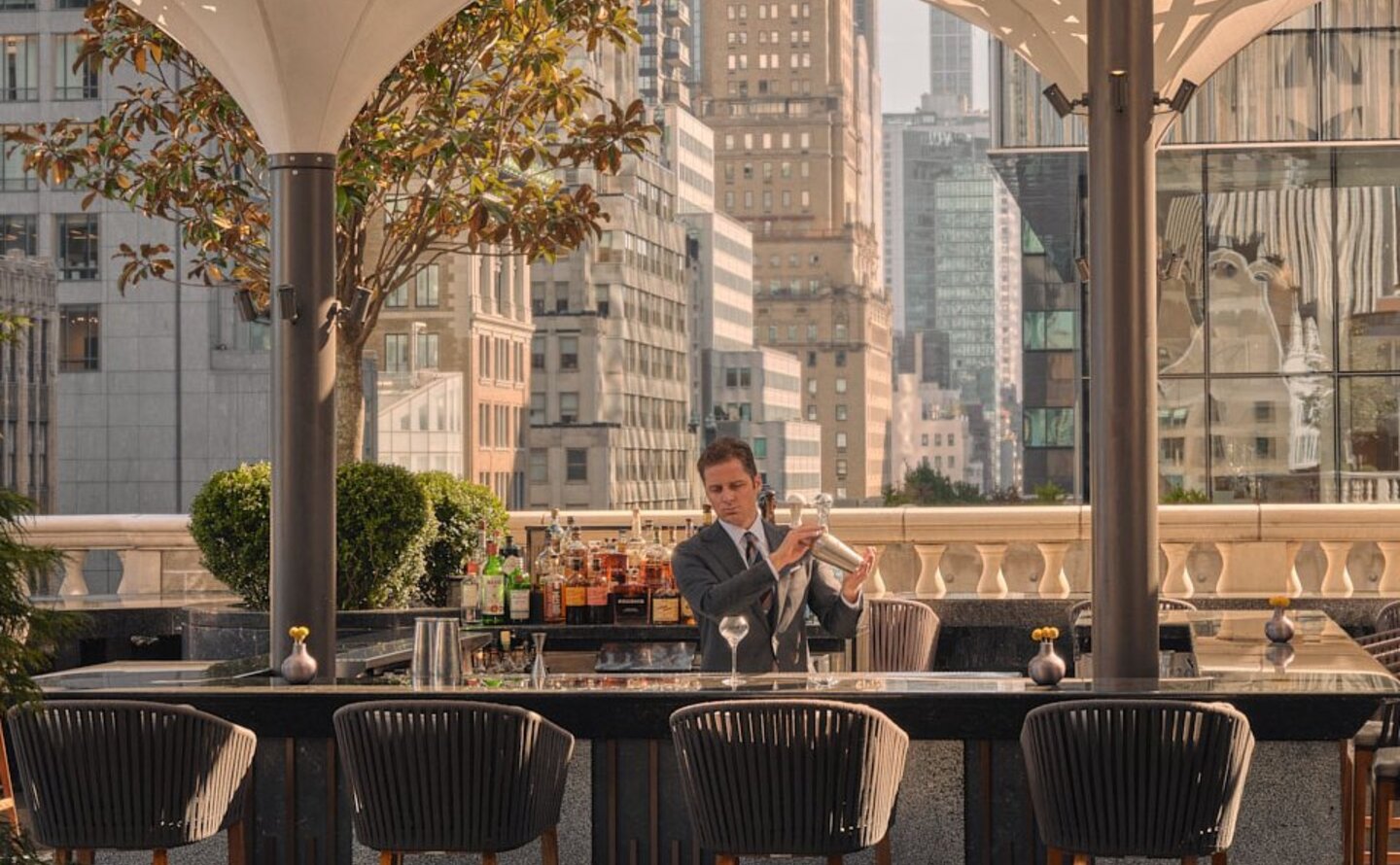 Bartender crafting cocktails at Aman New York's corner bar and garden terrace, shaded by canvas awnings.