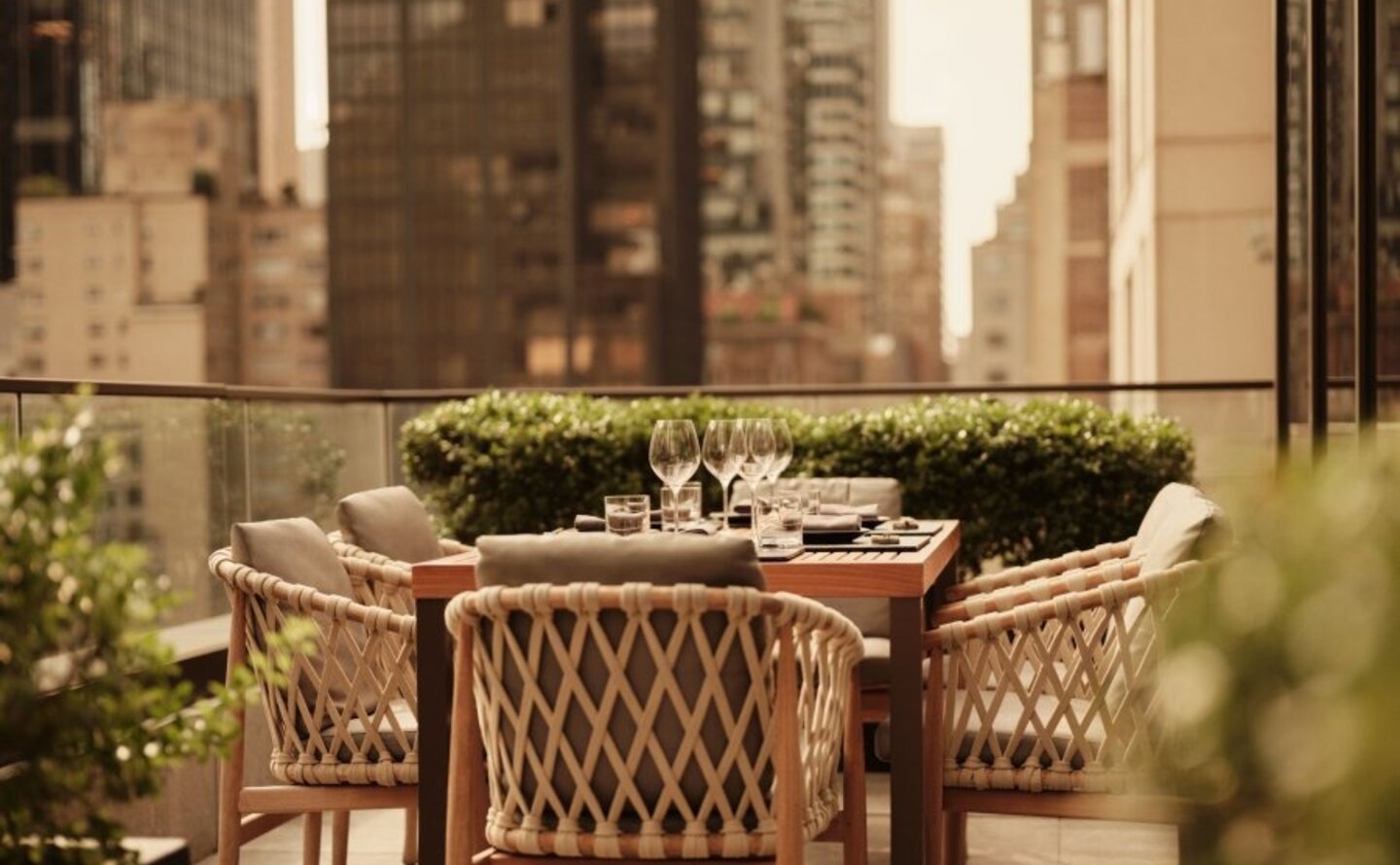 Woven chair and table on a sunlit terrace at Aman New York, with city skyscrapers visible beyond.
