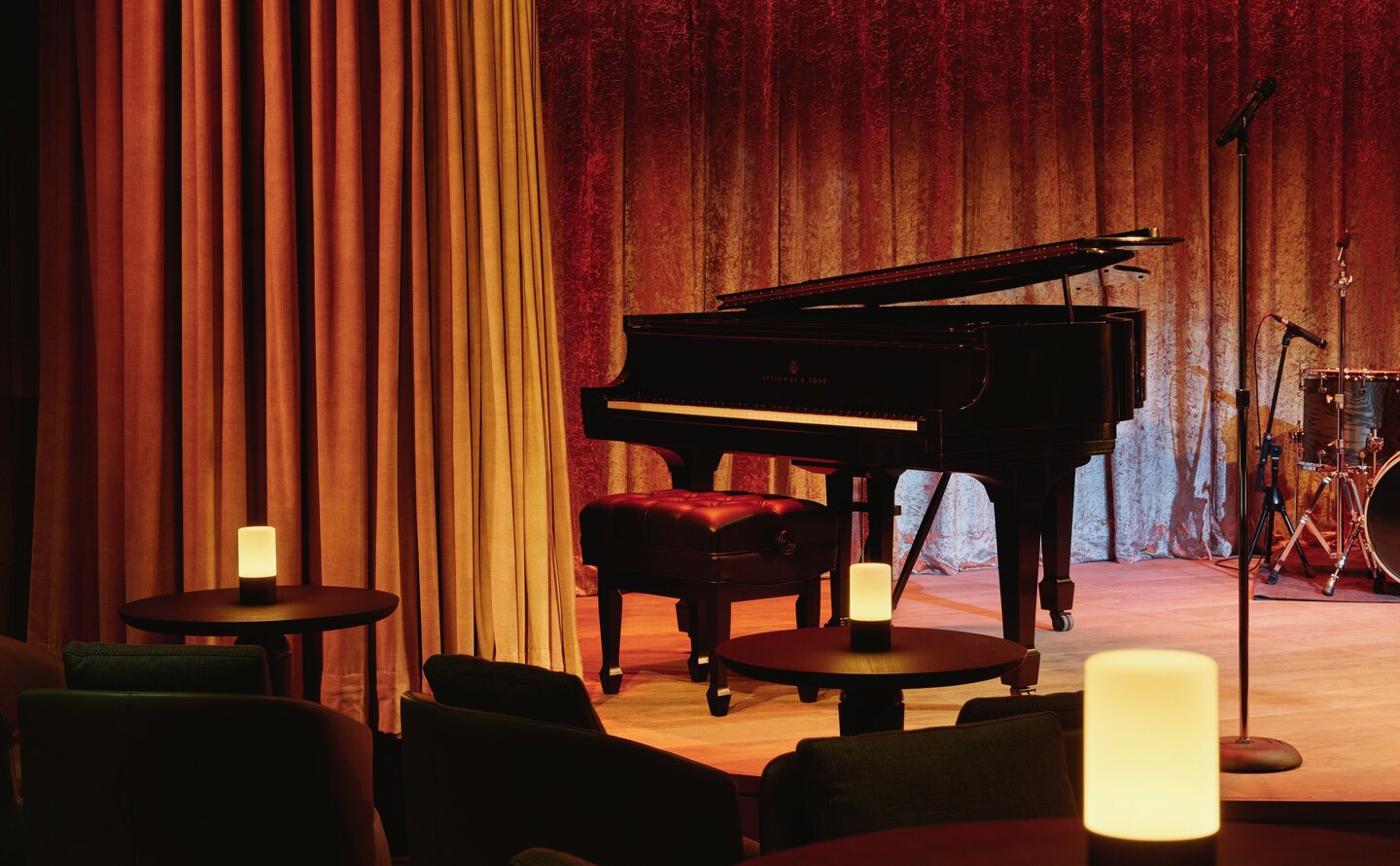 Grand piano on stage at Aman New York's jazz club, framed by warm red velvet curtains.