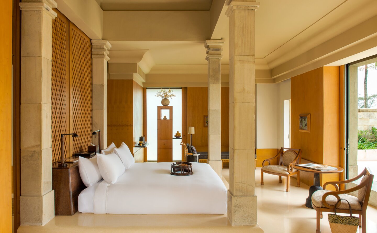 Amanjiwo pavilion bedroom interior with cream walls, wooden screens, and seated area by window.