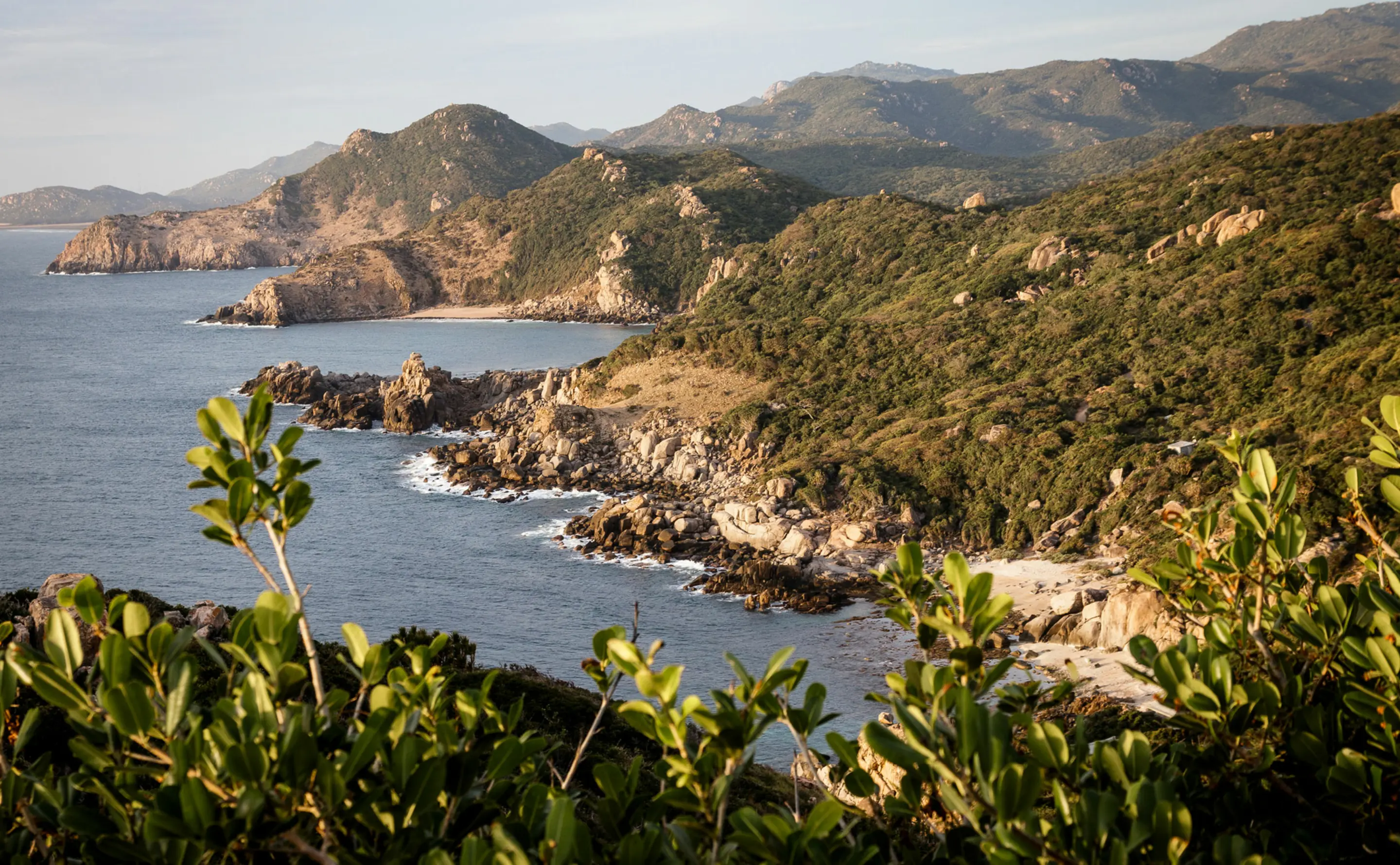 The Vinh Hy shore in late afternoon light, granite headlands and a boulder-strewn spit dropping into deep blue water, a small pale crescent of sand tucked behind, and the scrubby green ridges of the national park stacking away to the north.