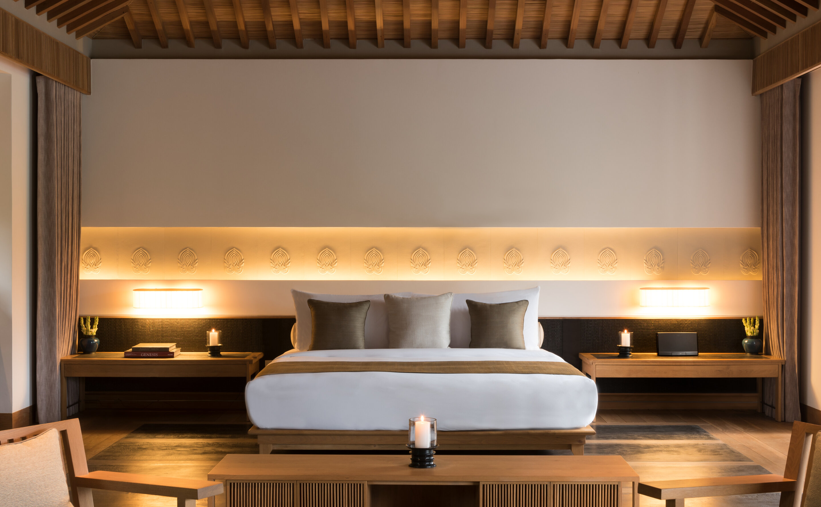 A pavilion bedroom under an exposed ceiling of close-set timber rafters, a low platform bed dressed in white and bronze between two long timber consoles, and a lit recess behind the headboard carved with a repeating lotus medallion.