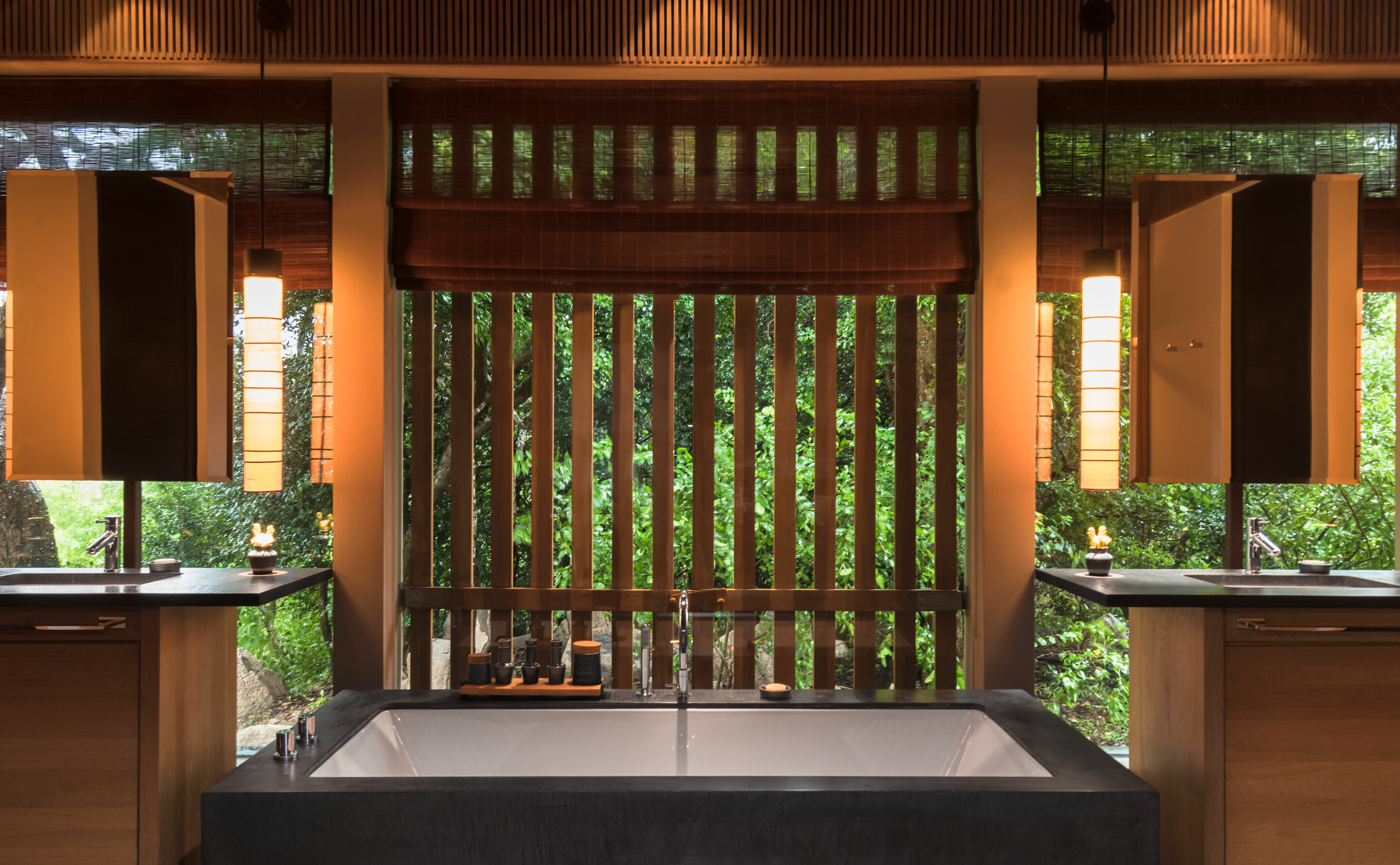 A pavilion bathroom built symmetrically around a dark stone tub, twin timber vanities with black stone tops and paper-shaded pendants either side, and a wall of vertical timber louvres with a raised roman blind opening straight onto green forest.