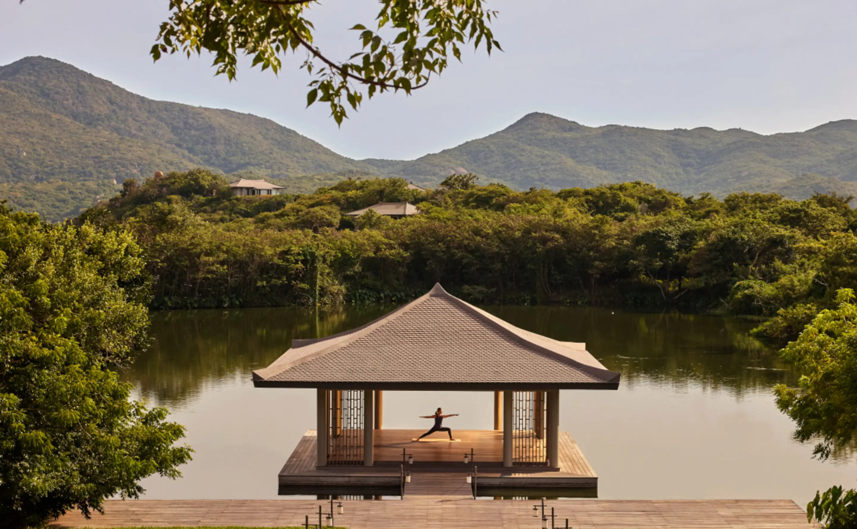 The yoga pavilion standing on its own timber platform out in the lotus lake, an open-sided pagoda with a broad tiled roof and lattice screens, a single figure holding warrior pose inside it, and forested hills folding away behind the far shore.