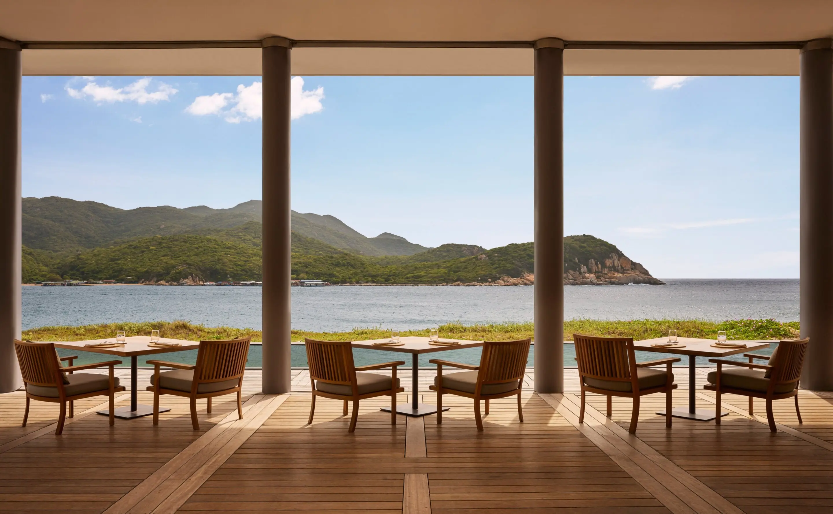The Beach Club restaurant open on three sides, teak tables and slat-back chairs on a wide timber floor between heavy square columns, looking past a strip of dune grass and a dark pool to the boulder headlands of Vinh Hy Bay.