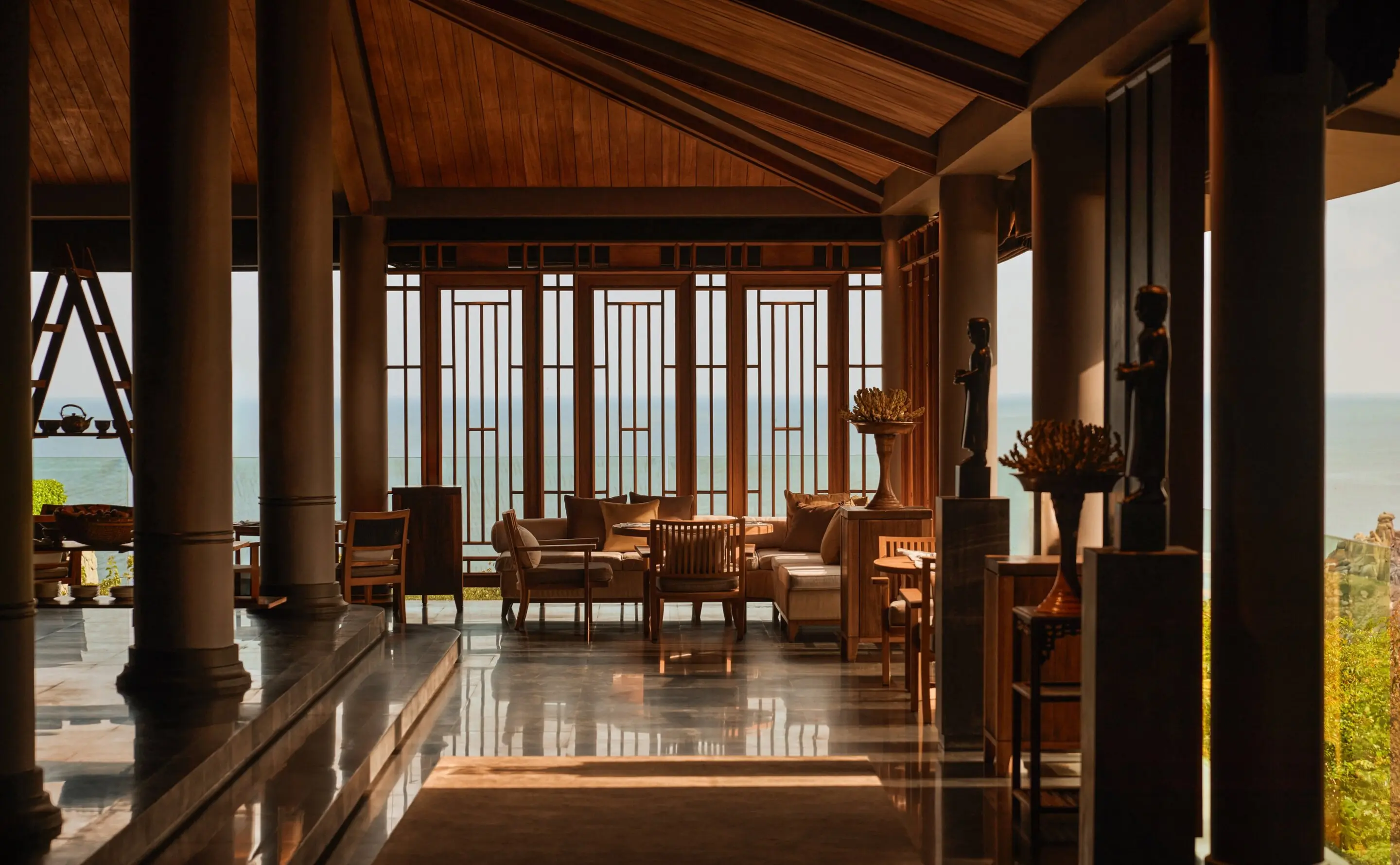 The main restaurant seen down its length: a polished stone floor between rows of pale columns under a pitched timber ceiling, low armchairs and dining tables set beneath tall lattice-panelled windows, carved wooden figures on plinths and the sea filling the openings on both sides.