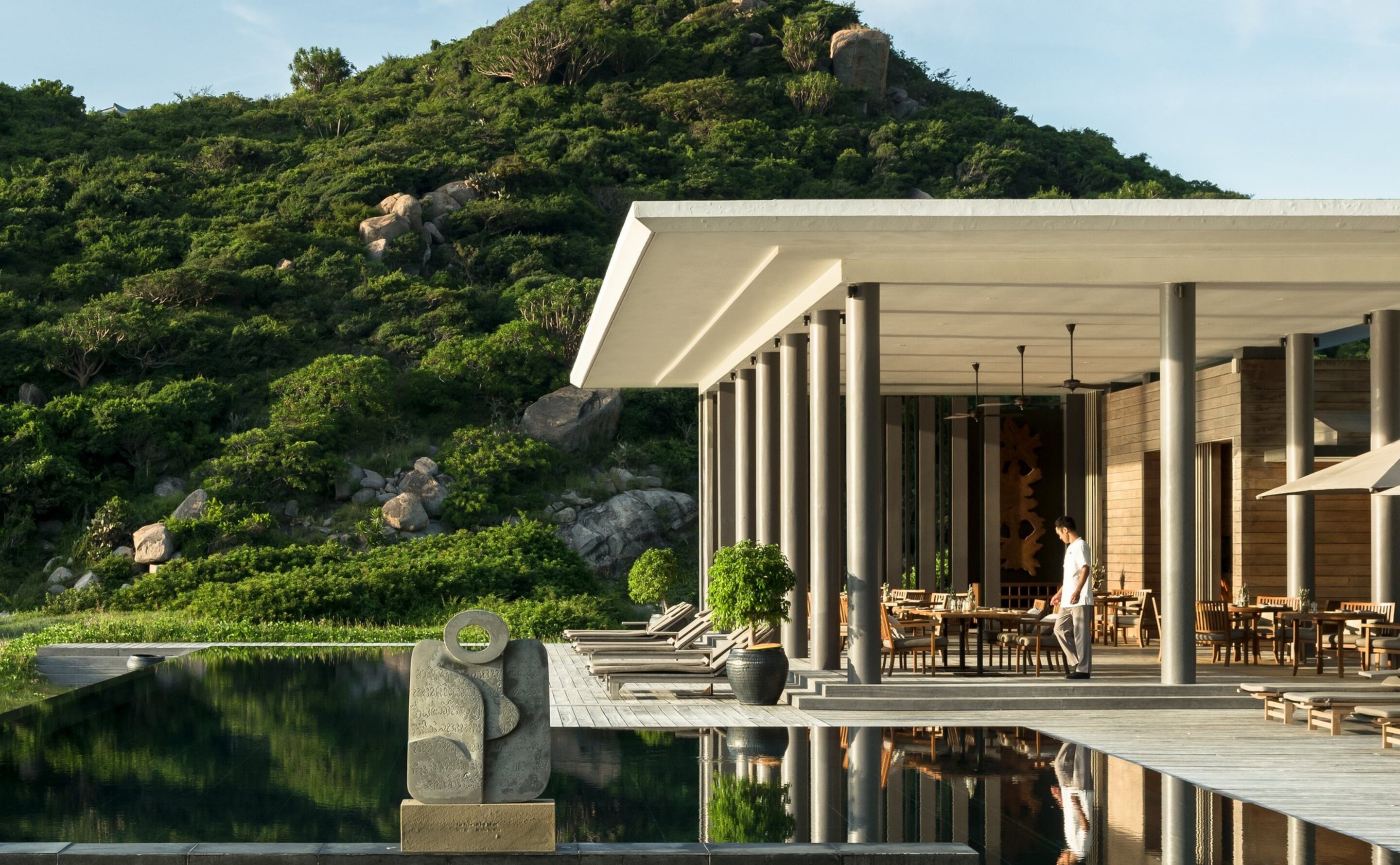 The Beach Club from across its black-water pool, a deep cantilevered canopy on slender pale columns shading loungers and dining tables, a carved stone sculpture on the pool's edge and a boulder-studded green hill rising straight behind.