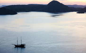 Sailing vessel anchored in calm waters at sunset, Amandira, Indonesia, with volcanic island silhouetted against twilight sky.