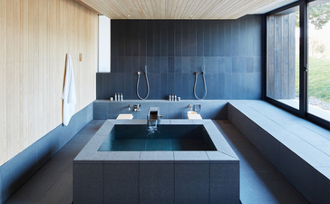 Baño minimalista con bañera de inmersión profunda en Amanemu resort, paredes azul oscuro y luz natural.