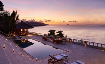 安缦丘澜度假村无边泳池沿海滨延伸，夕阳映照海面，金紫色天空倒映池水中。