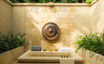 Borobudur Suite bathroom at Amanjiwo with sunlit stone walls, potted plants, and wooden bowl detail.