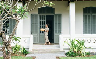 A guest stands in the doorway of a white colonial-style suite at Amantaka, framed by lush greenery and traditional architecture.